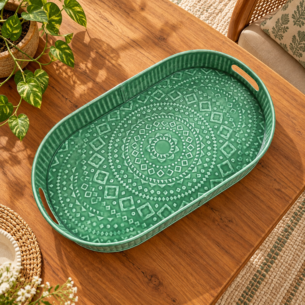 An oval green tray with a raised mandala pattern and cutout handles, shown on a wooden table with decorative plants and woven accents.