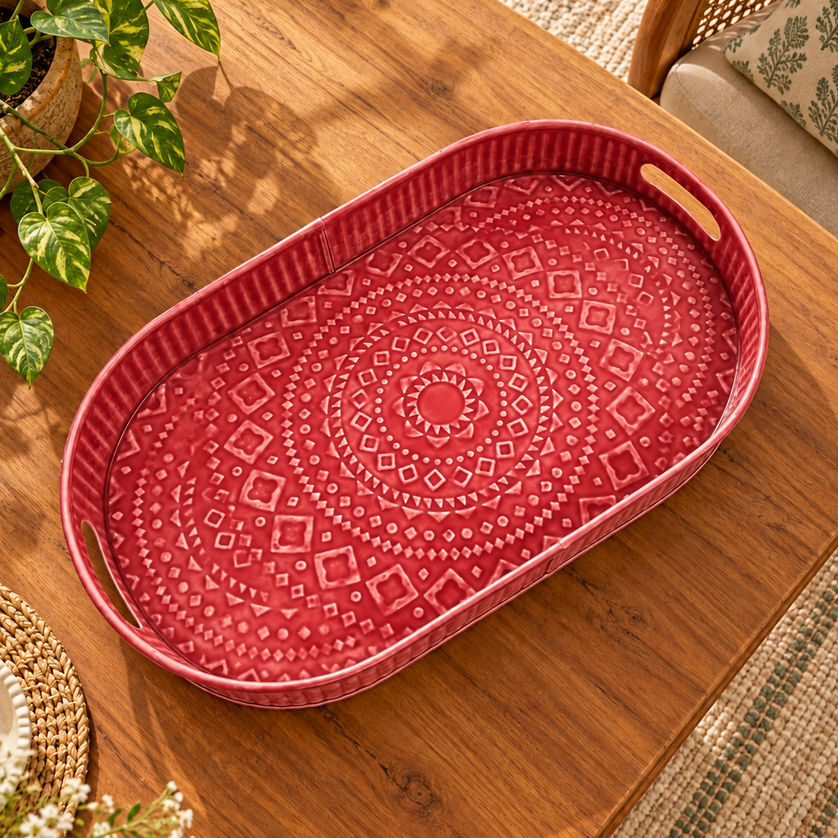 An oval red tray with a raised geometric mandala pattern on the bottom and light wood handles on the sides, shown on a wooden table with a plant in the background.